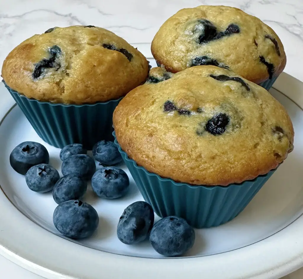 Three blueberry muffins on a plate with fresh blueberries