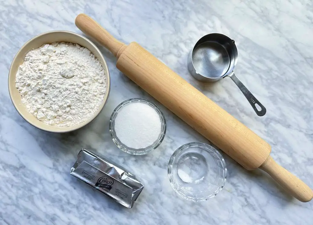 Individual bowls of flour, sugar, and salt next to vegetable shortening and a rolling pin on a white marble surface