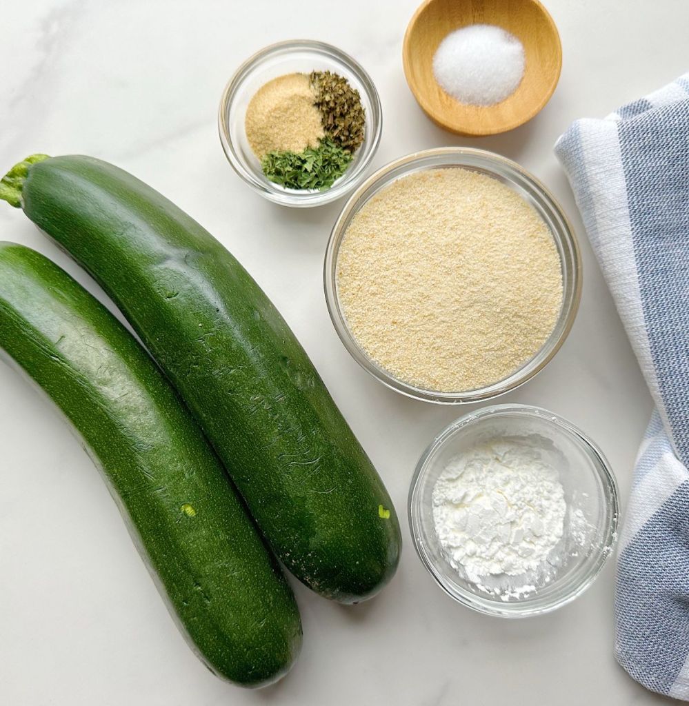 zucchini, breadcrumbs, spices and cornstarch on a white marble background. All ingredients needed for making air fryer zucchini fries.