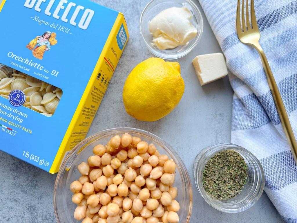 Chickpeas, lemon, herbs, box of pasta, and vegan cheese on a grey background