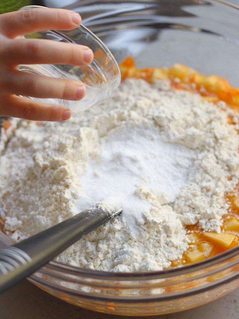 a tiny helper pouring dry ingredients into a large glass bowl for vegan carrot apple muffins